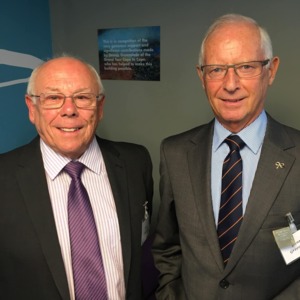 Phillip is standing to the left and Dennis is standing to the right. They are both looking at the camera and smiling. They are wearing suits and ties. 