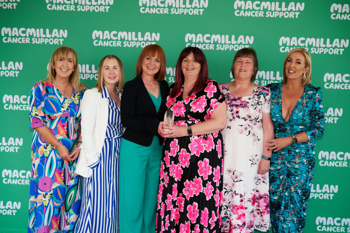 A group of women are standing and looking at the camera. In the middle two women are holding an clear glass award together. Behind them is a background with the green Macmillan logo on it.