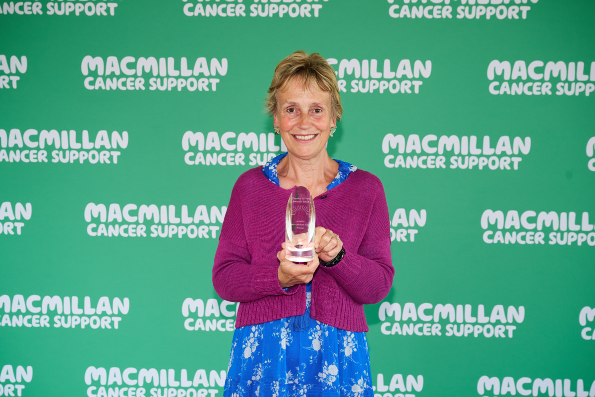 Jo is smiling at the camera in front of a green Macmillan background. She is wearing a purple jumper and blue patterned dress. She is holding a clear glass award in her hand.