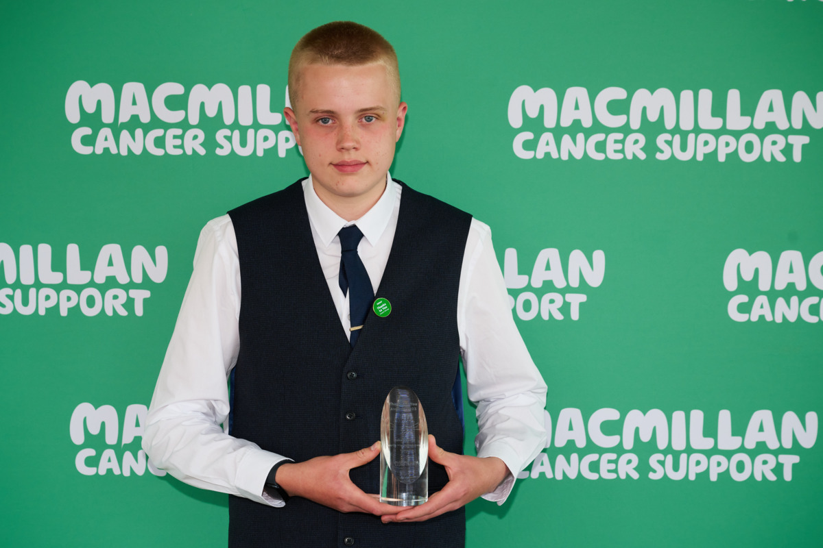 Jake is looking at the camera and behind him is a green background with the Macmillan logo on it. Jake is wearing a white shirt, black vest and black tie. He is holding a clear glass award in his hands. 
