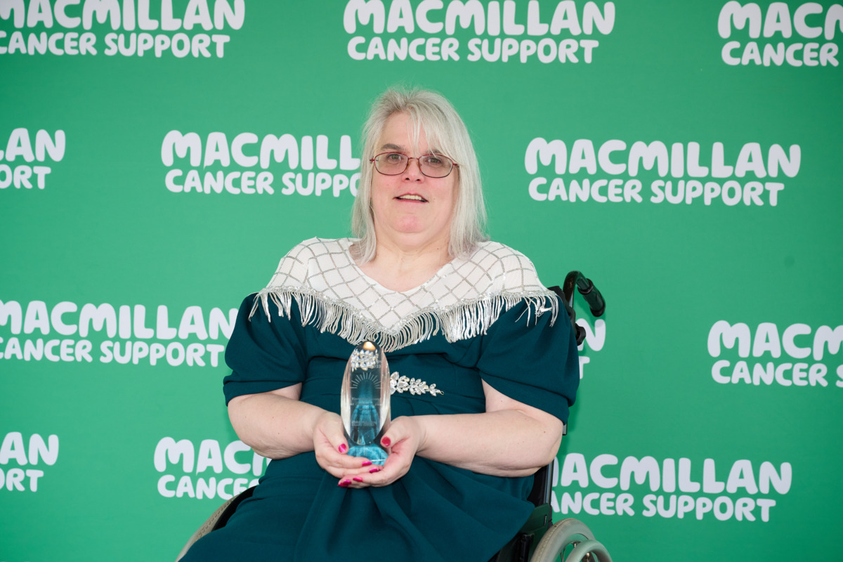 Fiona is looking at the camera. Behind her is a green background with Macmillan logos on it. She is wearing a blue dress with a white shall draped over her shoulders. 