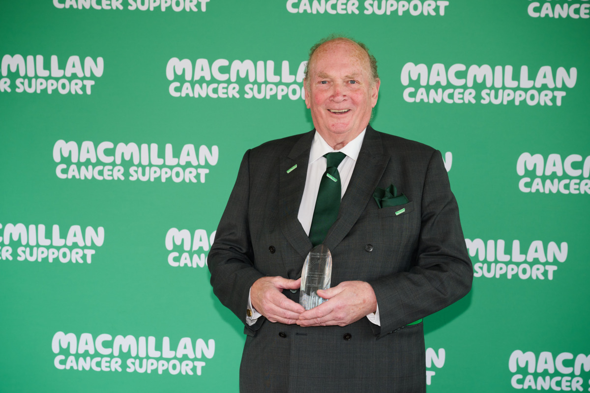 Brian is smiling at the camera. Behind him is a green background that has Macmillan logos on it. He is wearing a black suite with a green tie. 