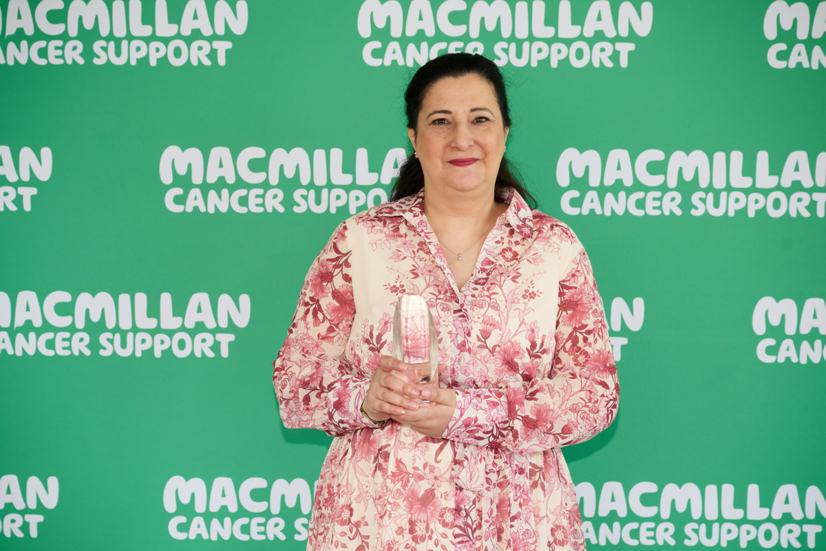 Ameena is smiling at the camera. Behind her is a green and white Macmillan background. She is wearing a red and white patterned dress. 