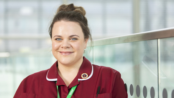 A headshot of Ria. She has brown hair that is tied up and she has blue eyes. She is wearing a red healthcare uniform and a green Macmillan lanyard.