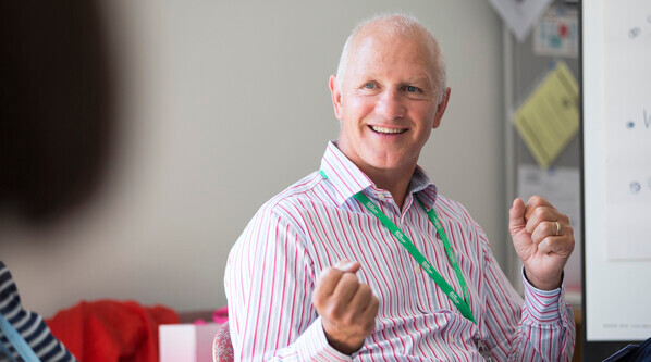 Paul is a Macmillan GP. He is sitting on a chair. He is wearing a colourful, striped shirt and green Macmillan lanyard. He appears to be caught in mid conversation.
