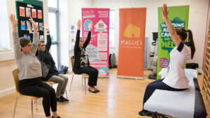 Karen Livingstone is a Macmillan Clinical Specialist in Physiotherapy and Lymphoedema. Karen is sitting on a hospital bed. Her arms are in the air. She has on a white NHS uniform and her long hair is pulled into a ponytail. Across from her are three patients sitting in chairs with their arms in the air. In the background are posters for different cancer services and support.