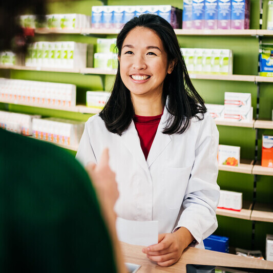 A pharmacist is speaking to a patient. The pharmacist has short black hair. They are wearing a red top underneath their uniform. They appear to be holding a sheet of paper.
