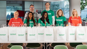 A group of individuals are standing behind a table which has white paper bags on it with the Macmillan logo. The individuals are wearing green Macmillan logo t-shirts or Santander logo t-shirts.