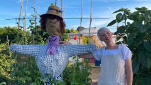 Lara is standing in a garden next to a scare crow. She is wearing a white and yellow striped dungarees. Her hair is cut short and is blonde.