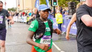 A woman is running in an event taking place on a road. She is wearing a blue Nike hat, a green Macmillan top, a black running vest, and orange running trousers. Behind her people are cheering at the event.
