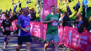 Two runners at an event. The person on the right is wearing a green Macmillan running top and shorts. Behind him are people cheering on event participants.