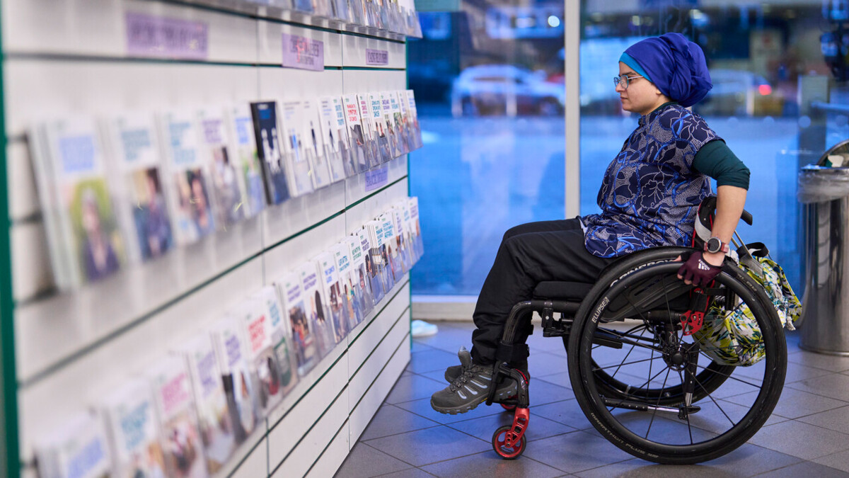 A woman is looking at cancer information booklets on display. She is wearing a blue headscarf and sitting in her wheelchair.