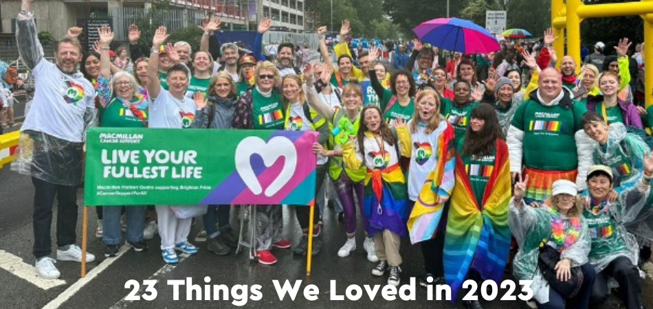 A large group of Macmillan volunteers wearing green Macmillan t-shirts and holding rainbow flags, waving at the camera and smiling.