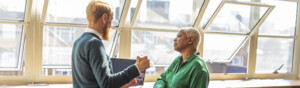 Two colleagues are standing near windows in an office and facing each other whilst talking. The person on the left is wearing a blue jumper and khaki trousers. The person on the right is wearing a green shirt and black trousers. They both are leaning on the window ledge.