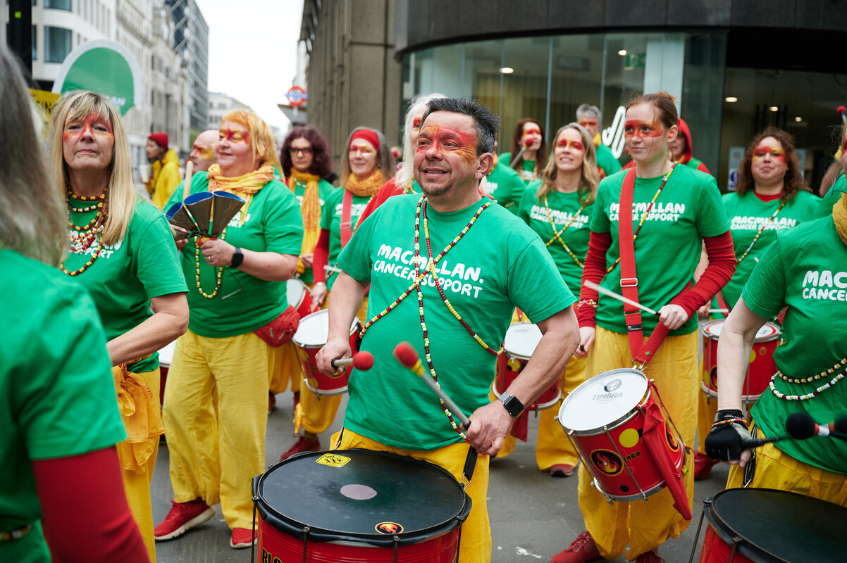A marching band are performing at the London Landmarks Half Marathon. Many of the performers having red face paint and are wearing green Macmillan tops and yellow trousers. They are playing different instruments like drums. 