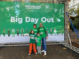 Marina and her family are standing outside in front of a large banner that has the words Big Day Out on it. Marina is pictured with her husband and 2 children. They are all wearing green Macmillan tops.