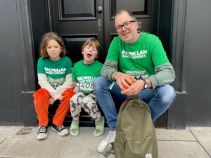 Marina's husband and two children are sitting on a door stump. They are all wearing green Macmillan tops. The door behind them is black and they appear outside.