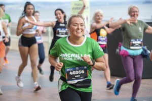 A woman in a green Macmillan top is running at an outdoor event. She has her blonde hair pulled back and is wearing ear buds. Pined to her top is an event number. Behind her are other runners.