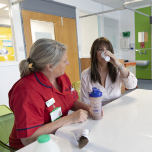 Lindsey Allen, a dietitian, is sitting with a patient in a hospital room. The patient is drinking from a plastic cup. Lindsey has a protein shake bottle in her hand. Lindsey is wearing a red NHS uniform. 