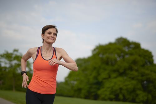Woman running outdoors. She has short brown hair and is wearing an orange vest.