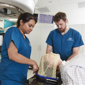 A person wearing a radiotherapy mask is lying down in a hospital room. Next to them are 2 radiographers who are examining the mask. 