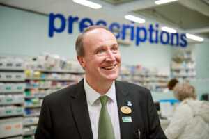 Graham Pickup is a Boots Macmillan Information Pharmacist. He is wearing a suit with a black coat and green patterned tie. Graham is smiling and has short brown hair and blue eyes. Behind him is a pharmacy with the sign prescriptions in large blue print. 