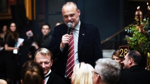 Comedian Al Murray is speaking into a microphone at a Macmillan Ball. He is standing near a group of people sitting down. Al is wearing a dark suit with a red and white stripped tie. He has short hair and a beard.