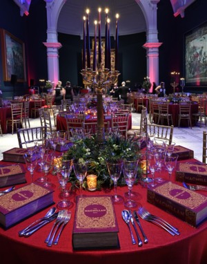 A table at a Macmillan ball. It has a candelabra in the centre with a green wreath and candles around it. There is a red table cloth on the table and glass and silverware. In the background are other tables set up in the same fashion.