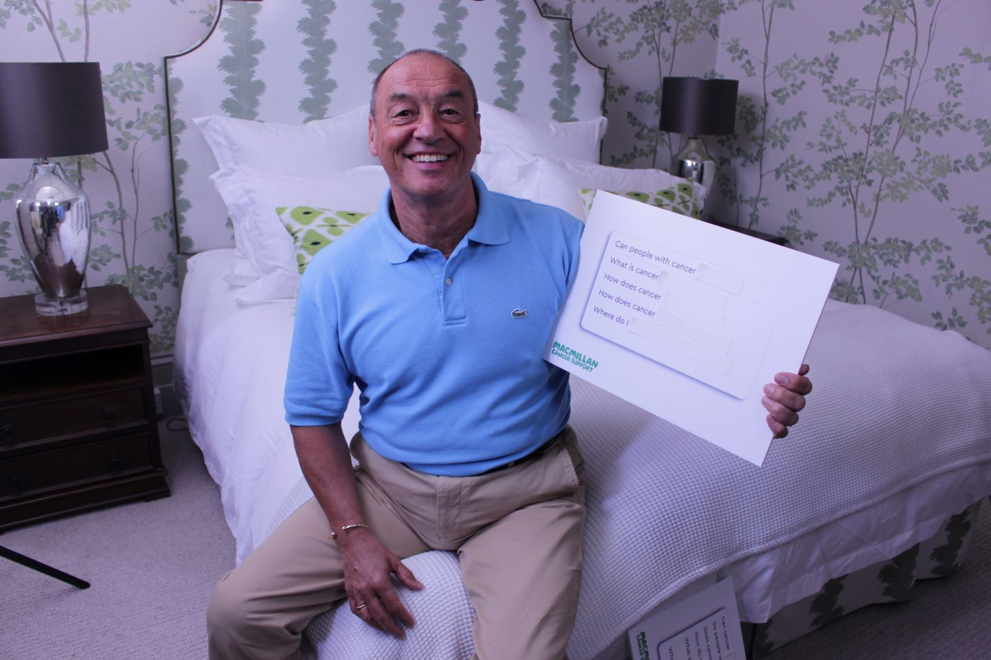 Elvin is sitting on a bed, looking at the camera and smiling. He is holiday a white board with questions about cancer written on it.