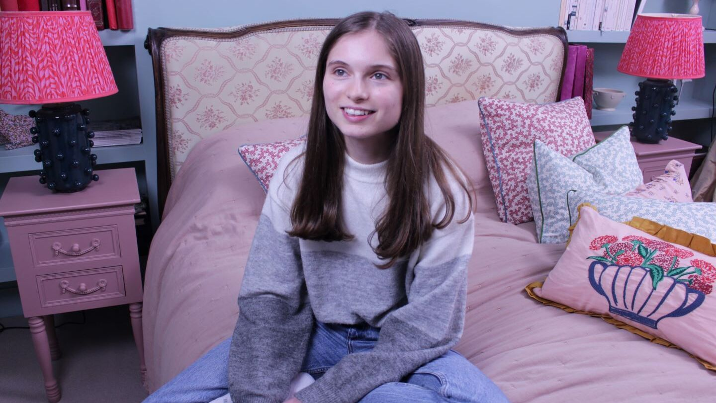 Ellie is sitting on a bed, looking up and smiling.