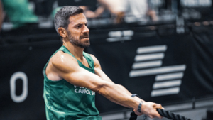 A participant is pulling at rope at a HYROX Event. They are wearing a green Macmillan top. They have short hair and are pulling a black rope. The background behind them is blurred.