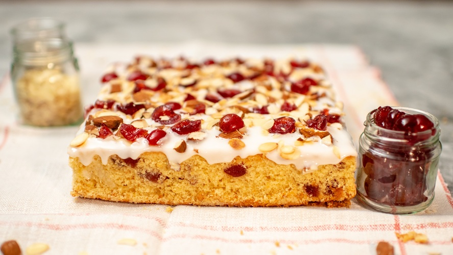 A golden-brown cherry and almond tray bake topped with smooth white icing, glistening glazed cherries, and sliced almonds. Presented on a striped cloth with jars of extra cherries and almonds beside it, 