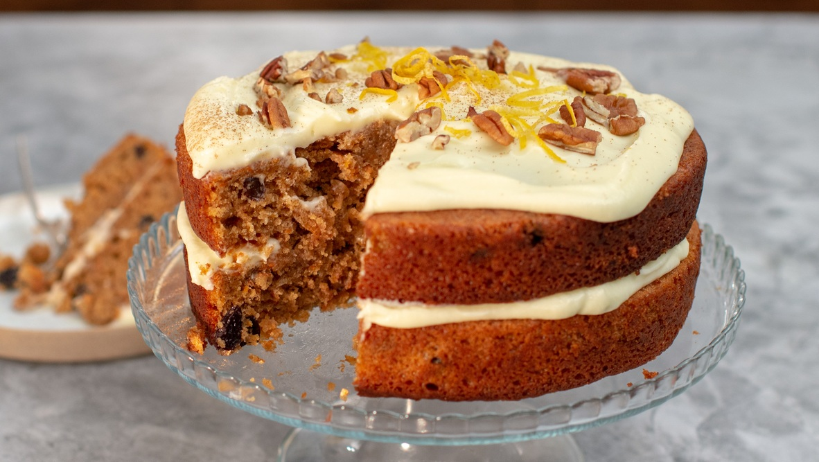 A moist, two-layer carrot cake with creamy white frosting between the layers and on top, garnished with chopped pecans and orange zest. A slice has been cut to reveal the rich interior speckled with carrot and raisins, all presented on a glass stand. 