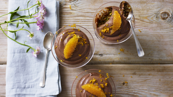 A view of vegan chocolate and orange cheesecake pots from above. The pots are placed on a wooden surface. They are decorate with an orange slice and zest. Next to the pots is a blue cotton napkin folded. On top of the napkin is a small spoon and some pink flowers. 