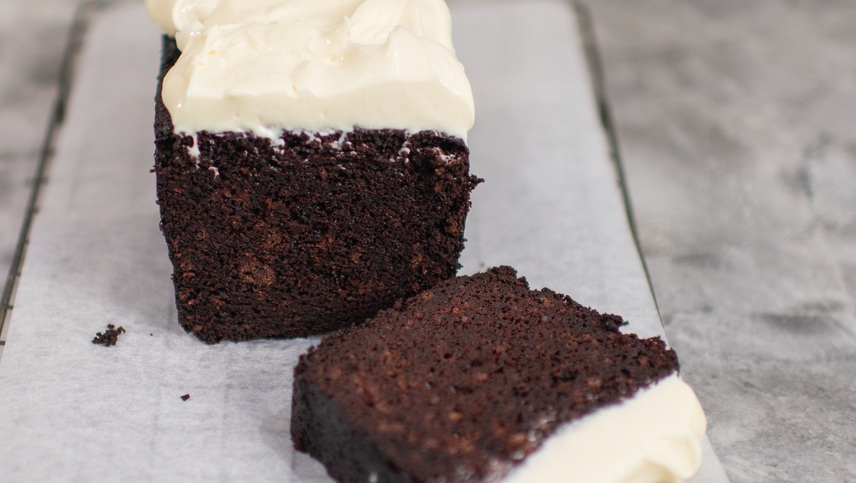 A rich, moist chocolate loaf cake topped with a thick layer of creamy white frosting, resting on parchment paper. A single slice lies in front, revealing its dense, decadent texture.