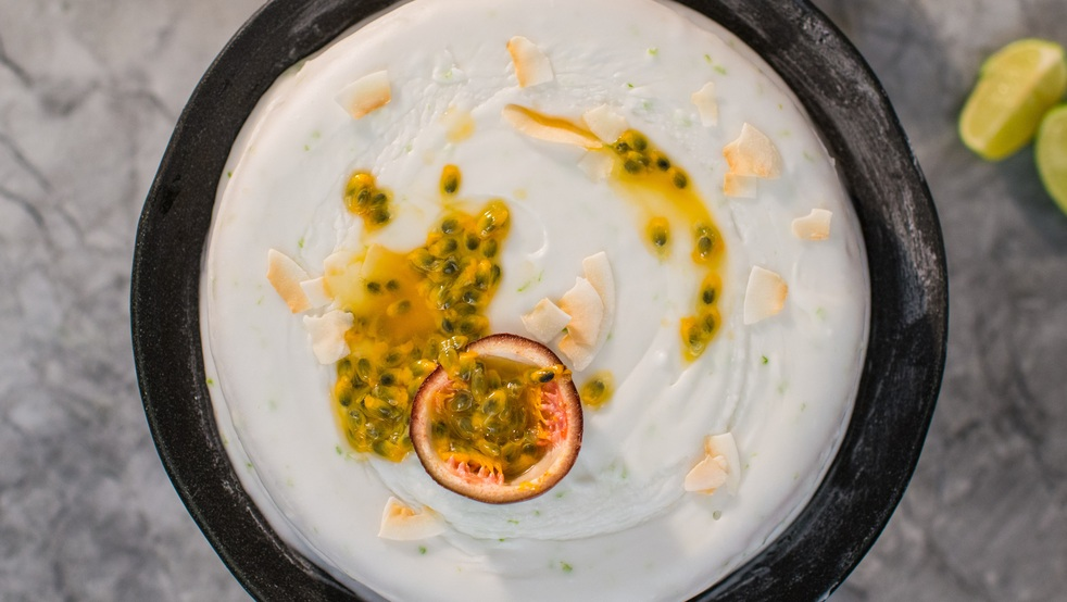 A view of a vegan lime, passionfruit and coconut cake from above. The cake is topped with a white frosting, half a passionfruit, and passionfruit seeds. It is in a dark rimmed container. 