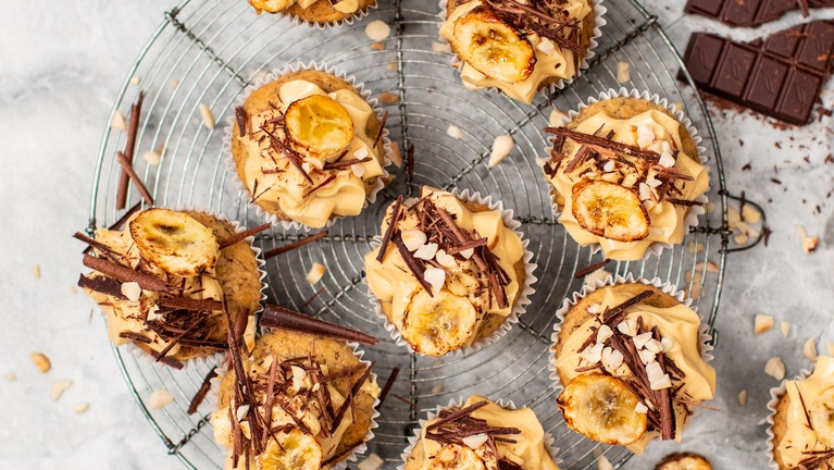 Several vegan peanut butter and banana cupcakes are on a cooling rack. The top of the cupcakes are decorated with frosting, chocolate flakes, and baked banana pieces. Next to the cupcakes is a broken bar of chocolate. 
