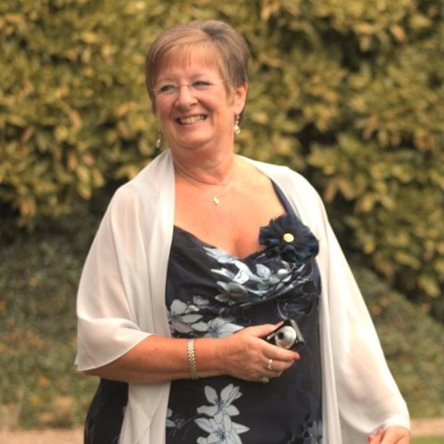 Mary is standing outside. She has short hair and is wearing a blue patterned dress and a white shawl. She is also wearing glasses. Mary is smiling and appears to be outside.