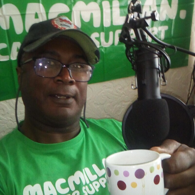 Damian is sitting near a radio microphone and holding a colourful mug. He is wearing a baseball hat and a Macmillan branded t-shirt. Behind him is a large Macmillan branded banner. Damian is also wearing black framed glasses.