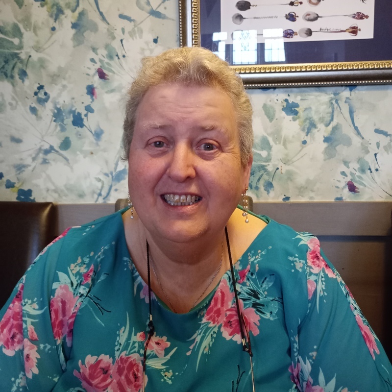 Vicky has short hair and is wearing a blue top with pink flowers. She is smiling. Behind her is a wall with floral wallpaper and a frame hung on the wall with artwork in it.