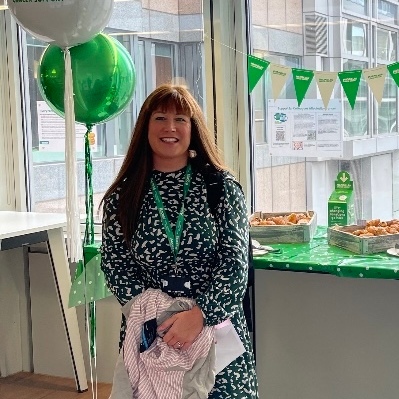 Louise has long brown hair and is wearing a black and white patterned dress. She is standing in a building that has Macmillan branded decorations. Behind her is a table with food on it and some balloons.