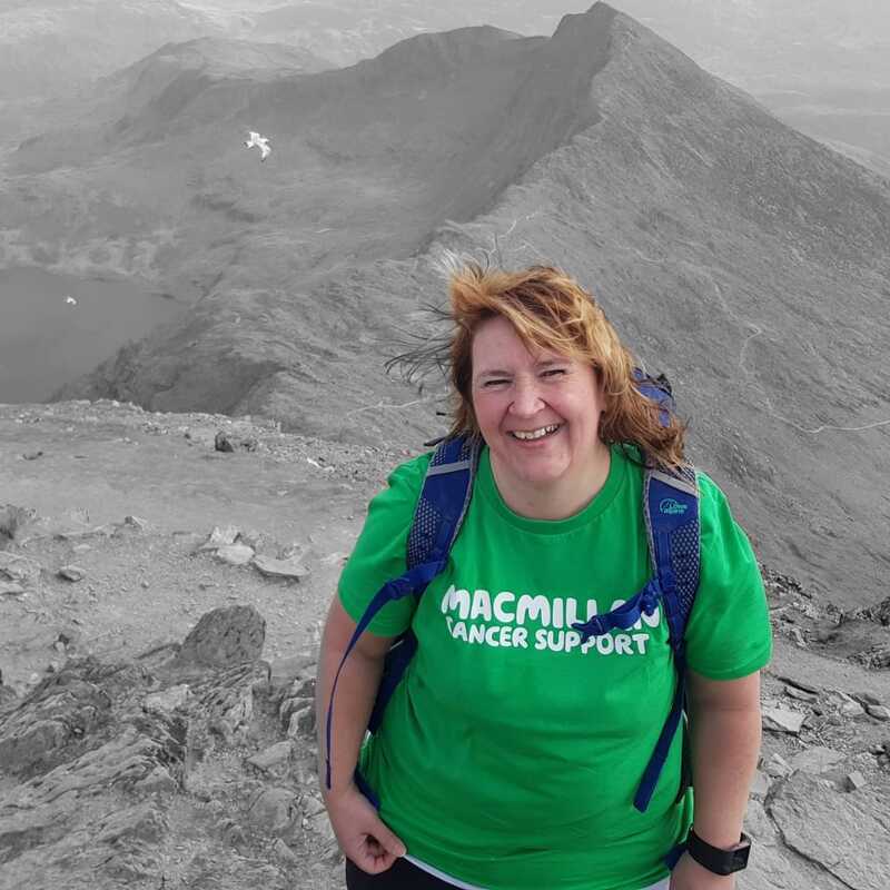 Sharon has short hair and is wearing a green Macmillan branded t-shirt. she is standing on top of a rocky mountain. The image has been altered so that Sharon is in colour but the rest of the photo is in greyscale.