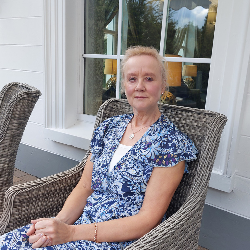 Geraldine is sitting on a wicker chair. She has her hair pulled back and is wearing a blue patterned dress. Behind her is a wall with a glass window.