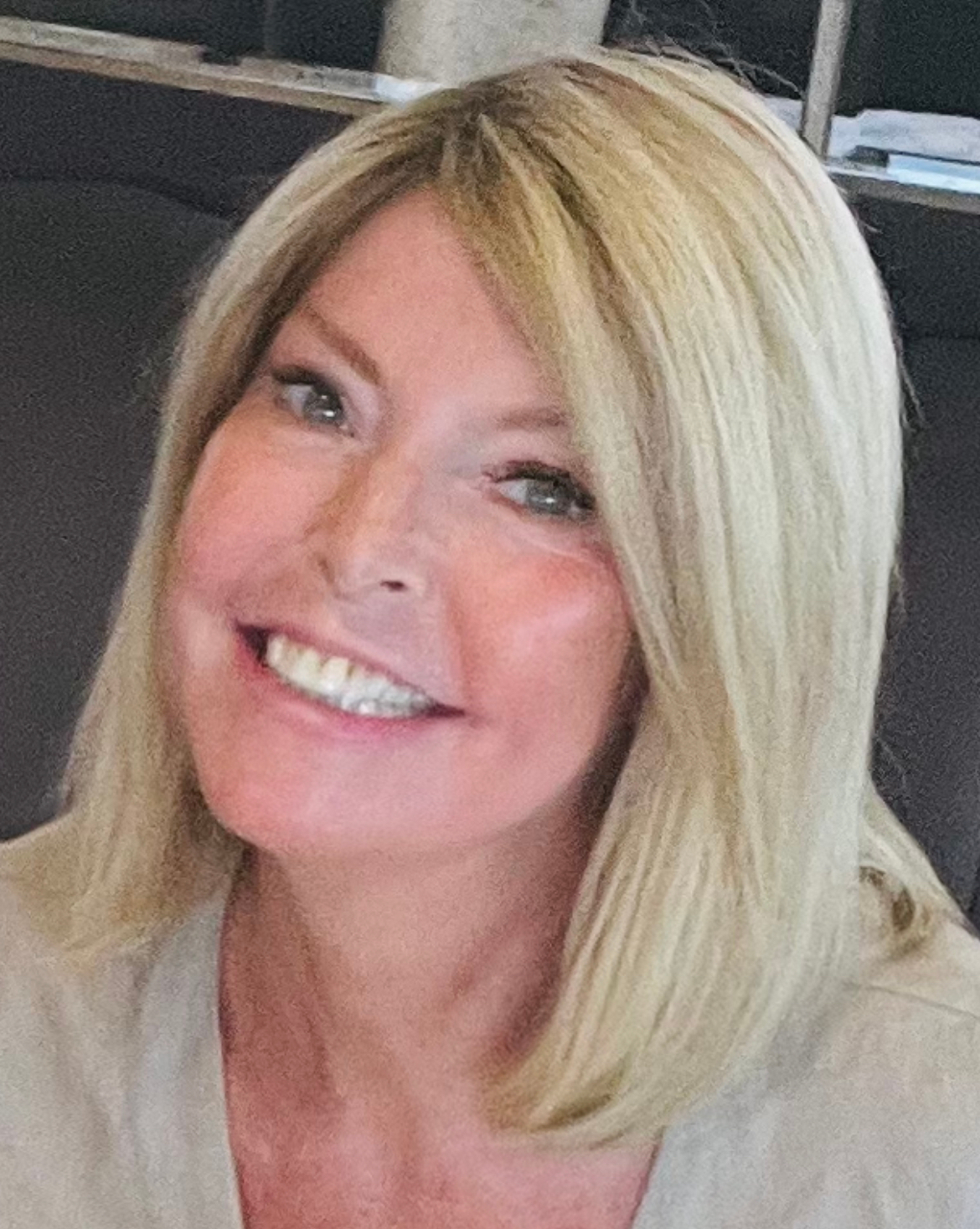 Carolyn has short shoulder length blonde hair. She is wearing a grey top and a yellow top underneath it.