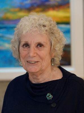 Penny has short curly hair. She is wearing a dark top that has a broach on it. Behind her is a colourful painting on a wall.