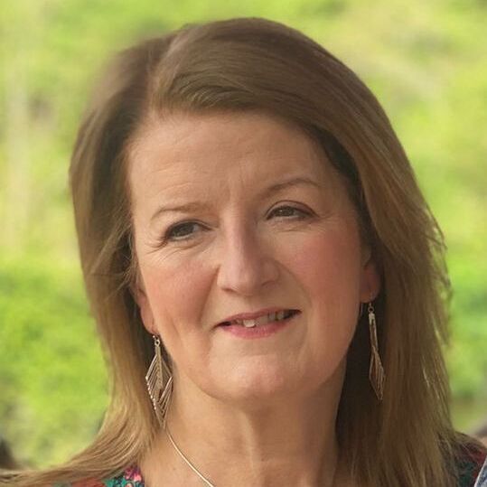 Ana has short shoulder length brown hair. She is wearing a colourful top and a necklace. The background behind her is blurred and green.