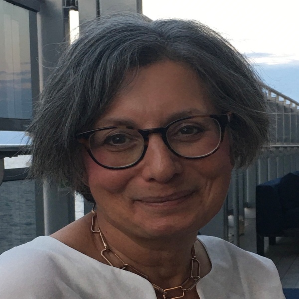 Nes has short dark hair and is wearing a white top and gold necklace. She is also wearing black rimmed glasses. She appears to be outside.