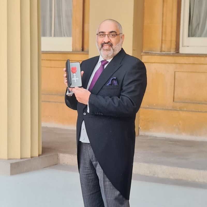 Salim is standing outside. He is wearing a full suit with a dark jacket, grey trousers, white shirt and a purple tie. He is holding a honorary award in his hands. He is also wearing dark rimmed glasses. 