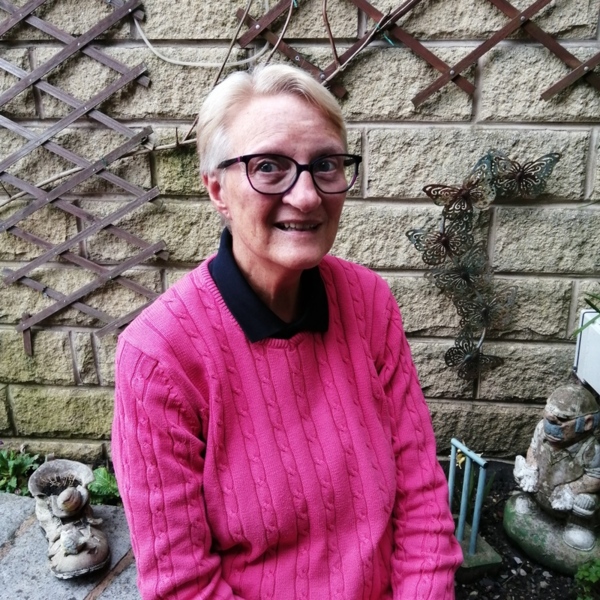 Kay has short hair and is wearing a bright pink jumper. She is also wearing a blue polo top. She appears to be sitting outside in a garden.