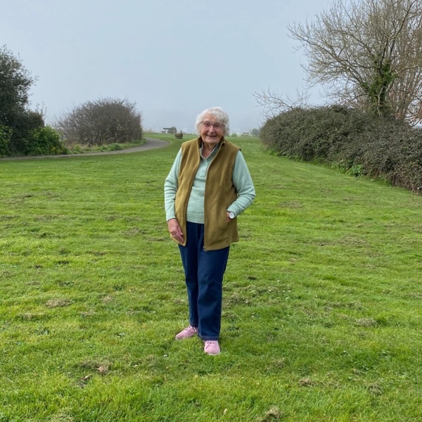 June is standing outside on a patch of green grass. She has short hair. June is wearing a blue top, dark blue trousers, and a brown vest.